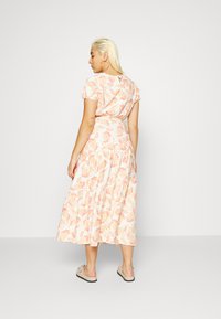Vestido maxi com padrão floral em pêssego claro e creme, com mangas curtas, cintura ajustada e saia em camadas. Combinado com sapatos bege tipo slip-on.