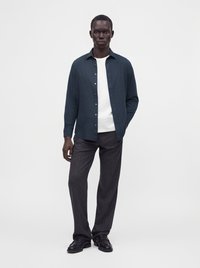 Homme portant une chemise bleu foncé boutonnée sur un t-shirt blanc, un pantalon gris foncé et des chaussures noires, debout devant un fond clair uni.