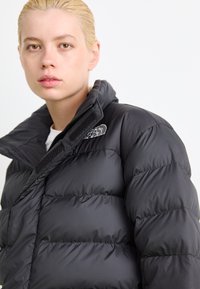 Schwarze Daunenjacke mit hohem Kragen, ausgestattet mit einem Reißverschluss und Klettverschluss. Der Stoff hat eine glatte Textur mit gesteppten Abschnitten.