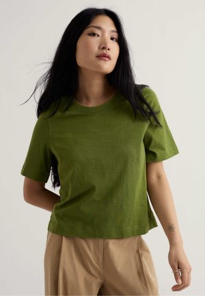 Vrouw met lang donker haar, gekleed in een groen shirt met korte mouwen en beige broek, staand met één arm achter haar rug.
