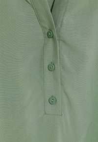 Koszula z krótkim rękawem, z kołnierzykiem, w jasnozielonym materiale o gładkiej strukturze, z trzema pasującymi guzikami z przodu i subtelnymi detalami w szwasach.