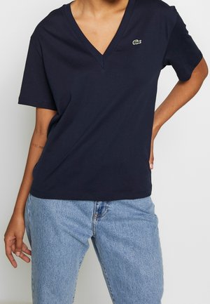 Persoon draagt een marineblauw V-hals T-shirt met korte mouwen met een klein groen krokodillenlogo en lichtblauwe hoge taille jeans.