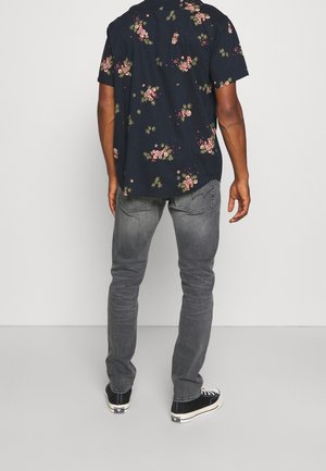 Camisa de flores de manga corta en color marino con flores blancas y rosas, combinada con jeans ajustados de color gris claro y zapatillas negras, vista desde la parte de atrás.