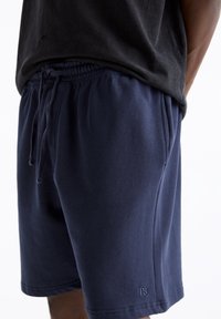 Shorts en coton bleu marine avec taille élastique et cordon de serrage. Présente un accent de logo subtil sur l'ourlet inférieur et une texture douce.