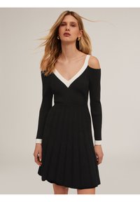 Robe noire en tricot côtelé avec des manches dénudées. Elle présente un col en V blanc et une jupe évasée. Manches longues avec des poignets blancs.