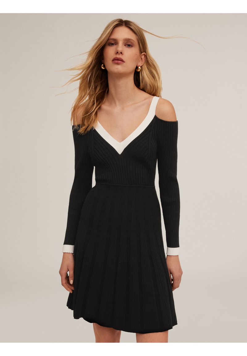 Robe noire en tricot côtelé avec des manches dénudées. Elle présente un col en V blanc et une jupe évasée. Manches longues avec des poignets blancs.