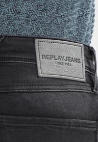 Jeans de mezclilla negra con una etiqueta de cintura de cuero gris texturizado que lleva la inscripción "REPLAYJEANS SINCE 1981" en lettering negro.