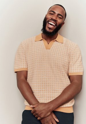 Kurzärmliges, v-ausschnitt Poloshirt in Hellorange mit einem strukturierten Muster und kontrastierendem Kragen. Modell lächelt mit verschränkten Armen.