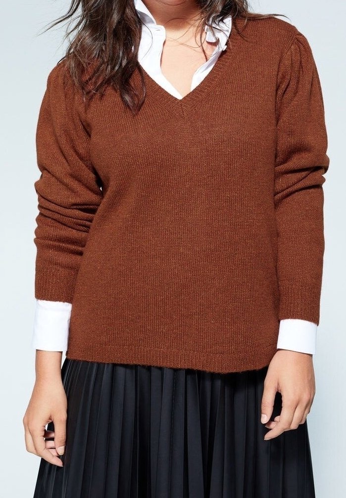 Femme portant un pull marron à col en V sur une chemise blanche à col avec une jupe plissée noire, debout devant un fond clair uni.