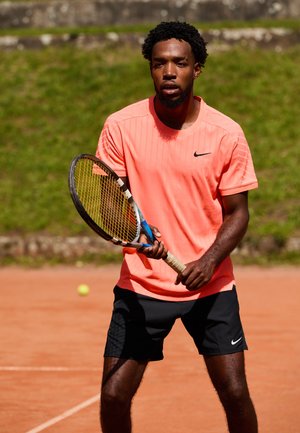 Joueur de tennis portant un tee-shirt corail vif de Nike et un short noir, tenant une raquette de tennis avec une poignée jaune, sur un court en terre battue.