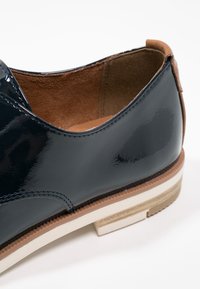 Chaussure habillée noire brillante avec doublure intérieure marron, semelle blanche, et détail de garniture marron sur le talon, vue en gros plan.