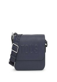 Bolso de piel azul marino cruzado con el logo "TOUS" en relieve, correa ajustable, herrajes plateados y textura suave. Forma rectangular.