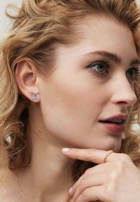 Srebrne kolczyki w kształcie motyli z misternymi detalami, na tle jasnych, kręconych włosów, obok złotego pierścionka na dłoni.