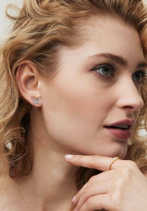 Srebrne kolczyki w kształcie motyli z misternymi detalami, na tle jasnych, kręconych włosów, obok złotego pierścionka na dłoni.