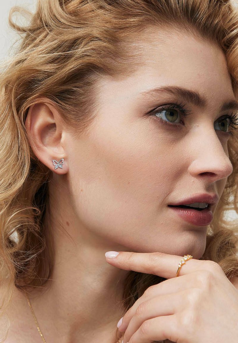Srebrne kolczyki w kształcie motyli z misternymi detalami, na tle jasnych, kręconych włosów, obok złotego pierścionka na dłoni.