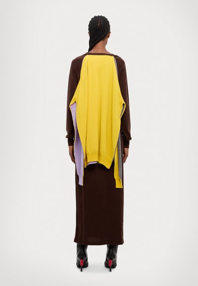 Robe longue marron avec un tissu drapé multicolore en jaune, gris et lavande dans le dos. Tissu texturé, longueur jusqu'aux chevilles, associée à des bottes à talons noires.