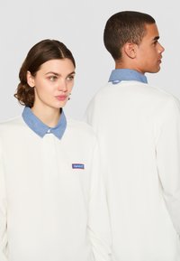 Camiseta polo blanca con un cuello de mezclilla, presenta un parche de logo rojo y azul en el pecho, mangas largas y una textura suave.