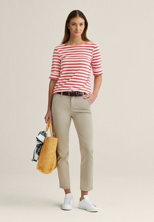 Nainen, jolla on punavalkoraidallinen paita, beiget housut, valkoiset lenkkarit, pitää käsissään kudottua ostoskassia, jossa on kuvioitu huivi, neutraalissa studiotilassa.