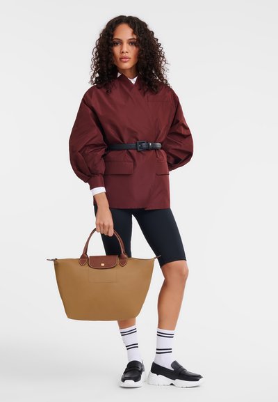 Brown tote bag with a textured surface and brown accents. Model wearing a burgundy oversized jacket, black bike shorts, and black slip-on shoes.