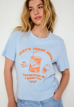 Frau mit welligem blondem Haar, die ein hellblaues T-Shirt mit einer Grafik eines Orangensaftkartons und Früchten trägt, dazu Text, der frischen Bio-Saft bewirbt.