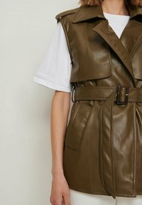 Zielona kamizelka z sztucznej skóry z dużym kołnierzem, paskiem w talii i kieszeniami z przodu. Nałożona na białą koszulkę. Gładka tekstura, strukturalny design.