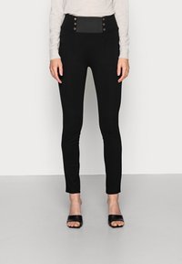 Pantalones negros de talle alto con una textura suave, detalles decorativos de botones y un corte ajustado, combinados con unas sandalias negras de punta abierta.