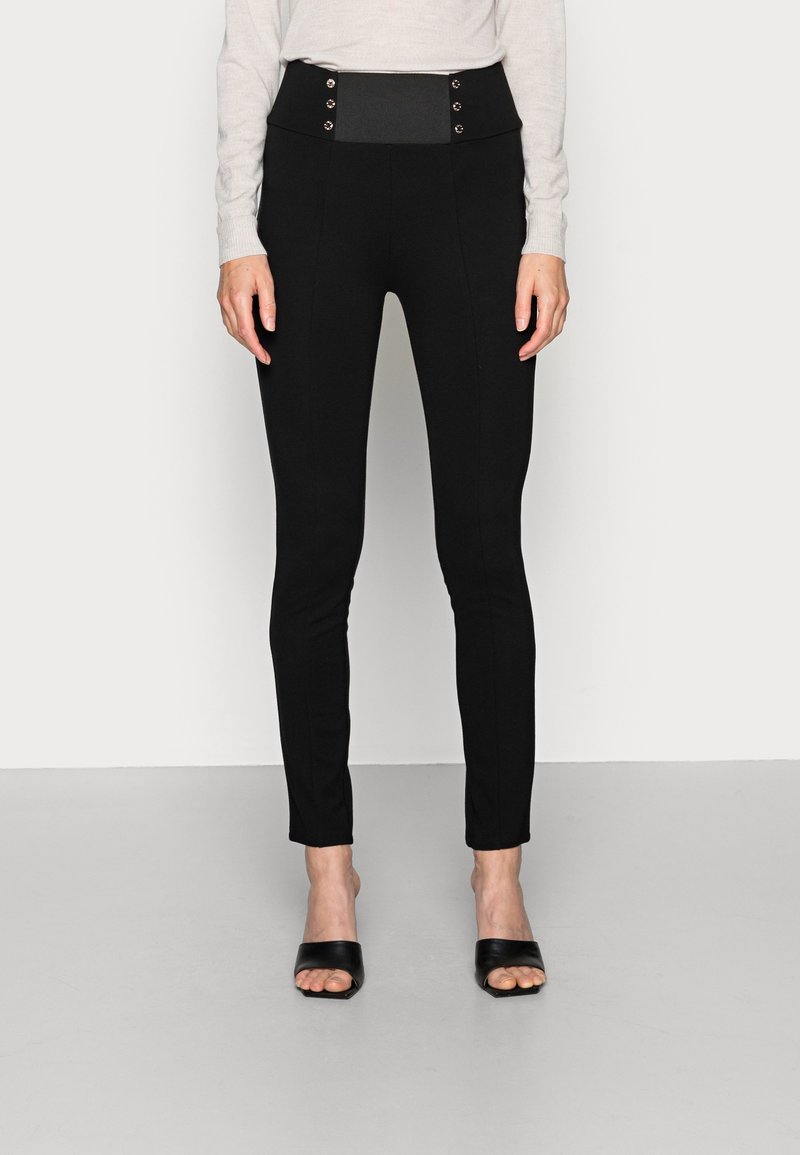 Pantaloni neri a vita alta con una texture liscia, dettagli decorativi con bottoni e una vestibilità slim, abbinati a sandali neri con apertura sulla punta.