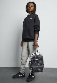 Otrok s dreadlocksi, oblečen v črn Nike kapuco, sivo Nike trenirko in črnima Nike supergami, drži črno-belo Nike nahrbtnik.