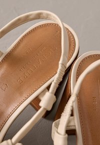 Sandales en cuir beige avec une semelle intérieure texturée marron et un talon en bois moyen. Elles présentent des brides tressées et des détails de marque en relief.
