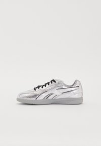 Srebrna metalna športna obutev z teksturiranim zaključkom, črnimi vezalkami in sivo podlogo. Ima črne akcentne stripe ob straneh.