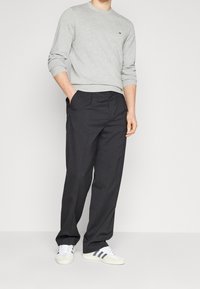 Uomo che indossa un maglione grigio chiaro, pantaloni neri larghi e sneakers bianche con strisce nere, in piedi con una mano in tasca.