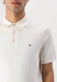 Muž v bílém polo tričku s krátkým rukávem, na kterém jsou tři knoflíky a malé červené, bílé a modré logo na hrudi.