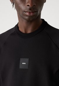 Μαύρη φούτερ μπλούζα με ραμμένο λαιμό και ράγλες μανίκια. Διαθέτει μια μικρή μαύρη ετικέτα με το "BOSS" σε ασημένια γράμματα στο στήθος.