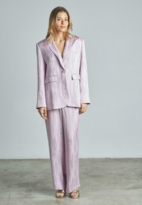 Suit lilla con tessuto testurizzato, caratterizzato da una giacca con rever ampio e pantaloni larghi. Include due tasche frontali, abbinati a sandali metallici.