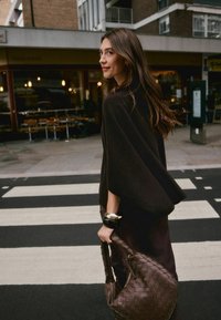 Brown poncho-style outerwear, smooth texture, paired with a dark brown woven handbag. Standing on a crosswalk with striped pavement.