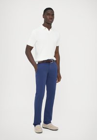 White short-sleeve polo with ribbed texture, paired with blue trousers and light-colored loafers. Brown woven belt adds detail.
