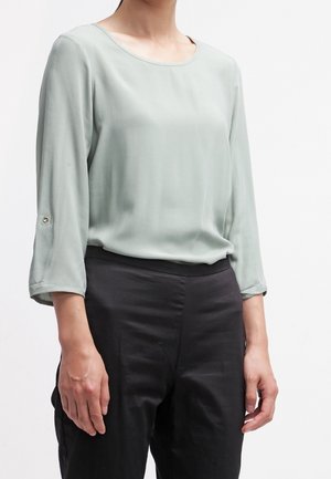 Kvinde iført lysegrøn bluse med lange ærmer og opklistrede manchetter samt sorte bukser med høj talje, stående mod en ensfarvet hvid baggrund.