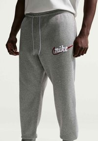 Bărbat purtând pantaloni de trening Nike gri cu șnururi albe și logo roșu brodat pe coapsa stângă, pe un fundal simplu.