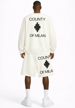 Mężczyzna w białej bluzie i spodenkach z czarnym napisem "COUNTY OF MILAN" i logo na plecach, w połączeniu z białymi skarpetami i sneakersami.