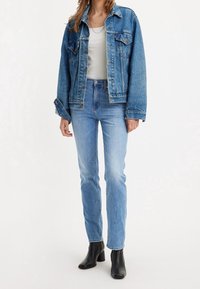 Chaqueta de mezclilla azul con un diseño clásico, que cuenta con dos bolsillos en el pecho, cierres de botones y un ajuste relajado. Combinada con jeans de un azul claro y botas negras hasta el tobillo.