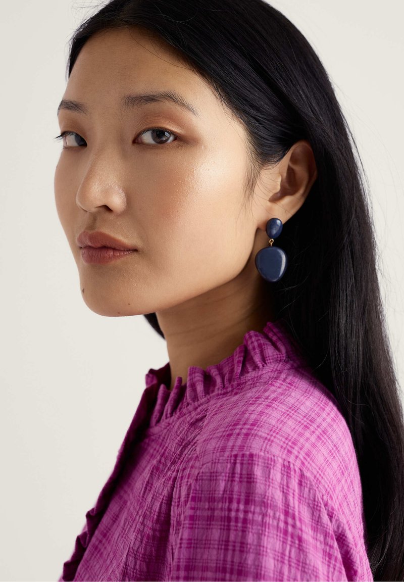 Boucles d'oreilles pendantes bleu marine avec un sommet rond et une forme en goutte. Le modèle porte un chemisier violet texturé avec un détail de col à volants.