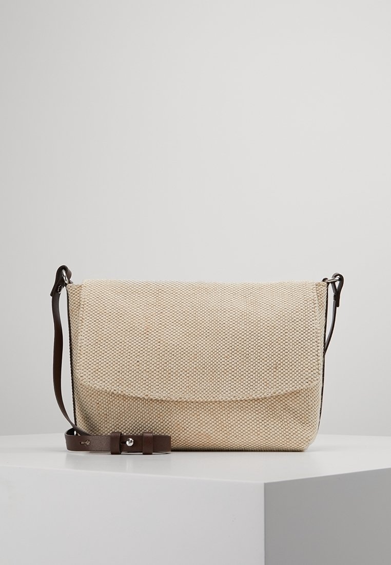 Beige vävd crossbody-väska med justerbart läderhandtag i mörkbrun som vilar på vit yta mot en enfärgad bakgrund.
