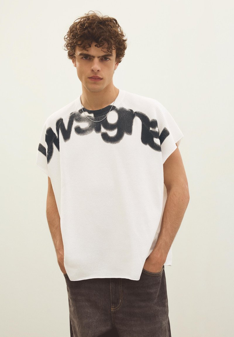 Jeune homme aux cheveux bouclés porte un débardeur blanc ample avec un texte graphique noir et un pantalon foncé, mains dans les poches, fond neutre.