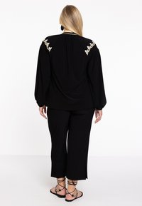 Zwarte outfit met een losse top met lange mouwen en geborduurde bloemen details op de schouders, gecombineerd met wijde zwarte broeken en sandalen met bandjes.