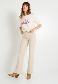 Camiseta crema con estampado púrpura "Ohlala"; combinada con pantalones acampanados beige claros y botines beige con tacón ancho.