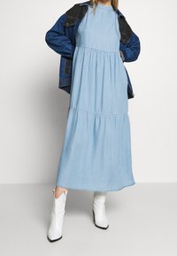 Lichtblauwe, mouwloze denim-maxi-jurk met een gelaagd ontwerp, gecombineerd met een donkere denimjack en witte western-stijl laarzen.