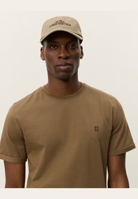 Casquette beige avec broderie "LOWER WEST SIDE" et un t-shirt à manches courtes marron arborant un petit logo rouge "II" sur la poitrine.