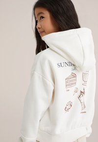 Felpa bianca con illustrazioni marroni di dolci e bevande sul retro. Realizzata in materiale morbido, con cappuccio regolabile e polsini a costine.