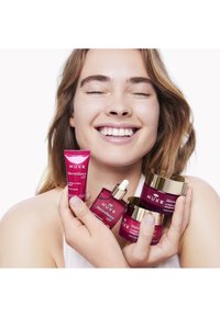 Productos de cuidado de la piel NUXE Merveillance Lift en tubos y frascos rosas con acentos dorados. Una persona sonriente sostiene varios productos cerca de su rostro.