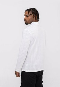 Chemise blanche à manches longues avec un motif texturé, col haut et coupe droite. Associée à un pantalon cargo sombre, présentée de profil.